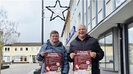 Annett Eine und Claus Roschanski stehen vor einem Gebäude und halten jeweils ein Plakat mit der Aufschrift '5. Jürgenohler Adventsmarkt'. Im Hintergrund ist eine beleuchtete Stern-Dekoration an einer Straßenlaterne zu sehen.