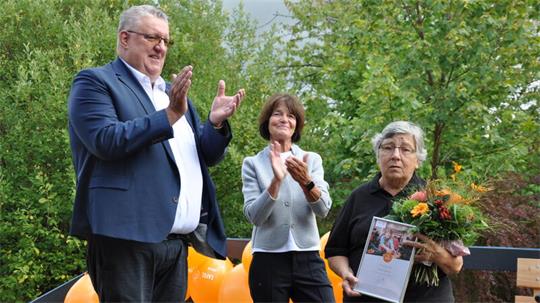 Drei Personen stehen vor grünen Büschen, zwei klatschen, eine hält einen Blumenstrauß und eine Urkunde