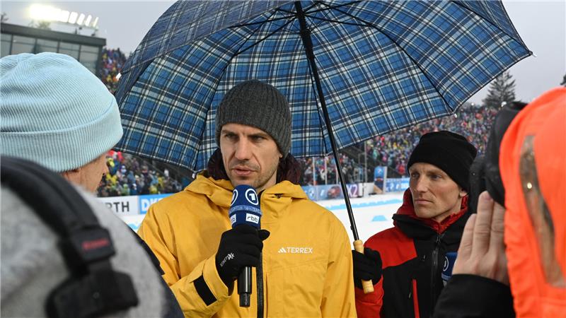 Zwei Personen mit Mikrofonen, eine trägt eine gelbe Jacke und hält einen blauen karierten Regenschirm, die andere eine rote Jacke, im Hintergrund eine Zuschauertribüne.