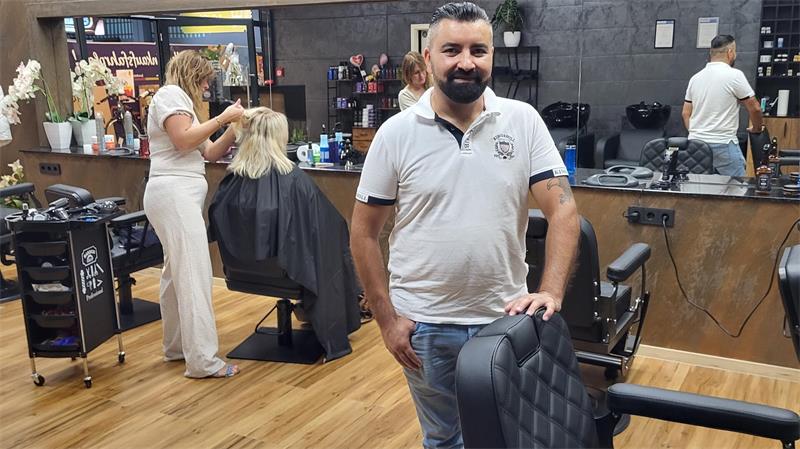 Mann mit Bart und weißem Poloshirt steht in einem Friseursalon und hält einen Friseurstuhl fest, im Hintergrund eine Friseurin bei der Arbeit