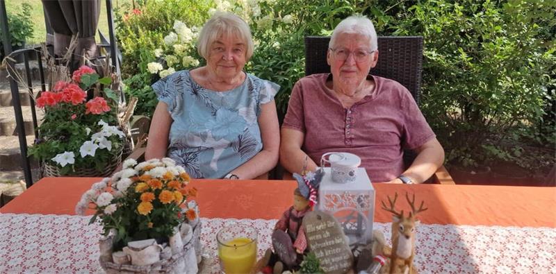 Zwei ältere Personen sitzen an einem mit orangefarbenem Tuch bedeckten Gartentisch mit Blumen und Dekorationen im Freien.