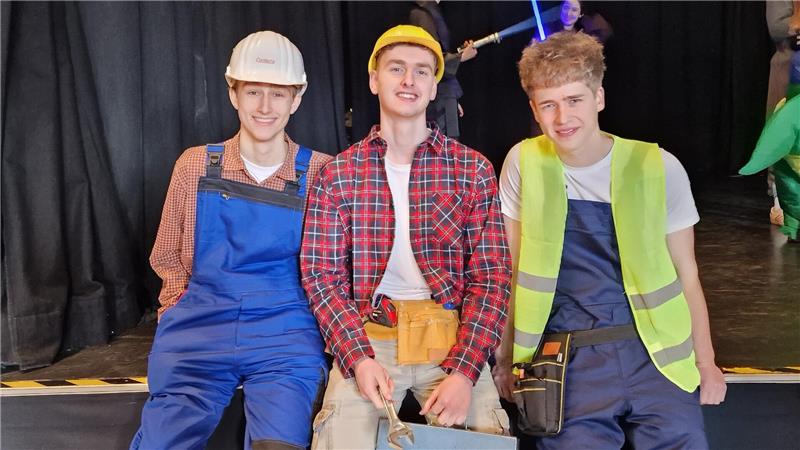 Drei Personen in Arbeitskleidung sitzen nebeneinander auf einer Bühne vor schwarzem Vorhang.