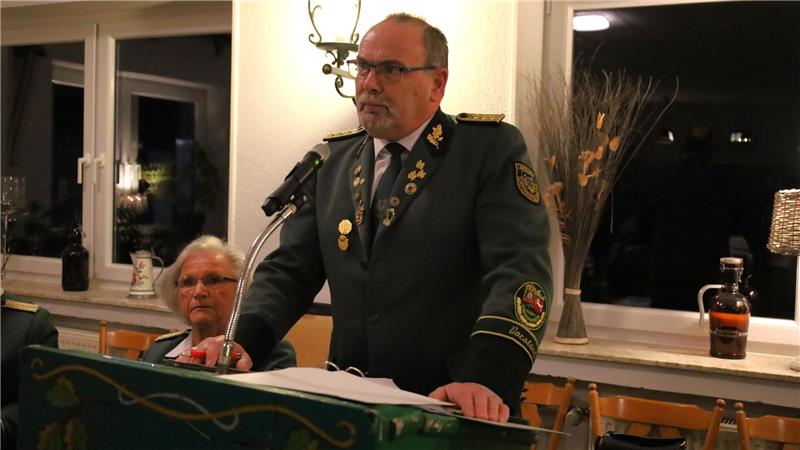 Mann in Uniform mit Abzeichen steht an einem Rednerpult in einem Raum mit Fenstern und Dekorationen
