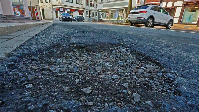 Nahaufnahme eines Schlaglochs in einer Asphaltstraße mit parkenden Autos und Gebäuden im Hintergrund.