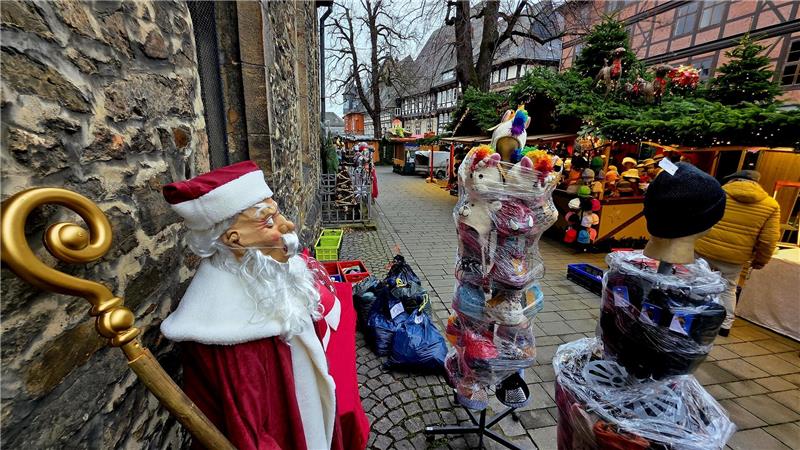 Weihnachtsmarkt Goslar: Öffnungszeiten, Parken und Co. Lebensgroße Nikolausfigur mit rotem Mantel und goldenem Stab lehnt an einer Steinmauer auf einem Kopfsteinpflasterweg mit Marktständen und zwei mit Folie umwickelten Schaufensterpuppen.