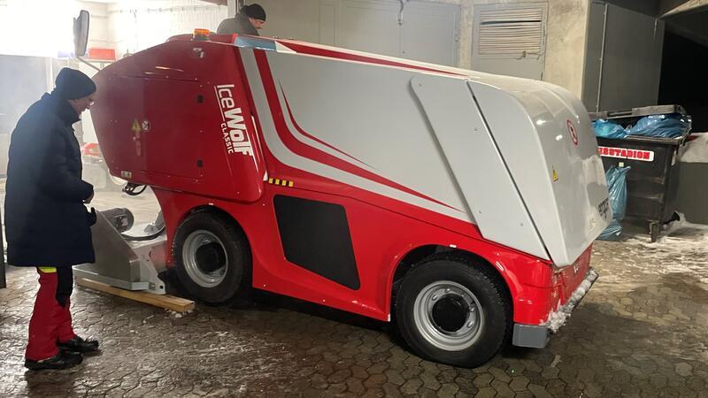 Rote und weiße Eismaschine der Marke "Ice Wolf" in einem Eisstadion, daneben steht eine Person in dunkler Jacke und roter Hose.