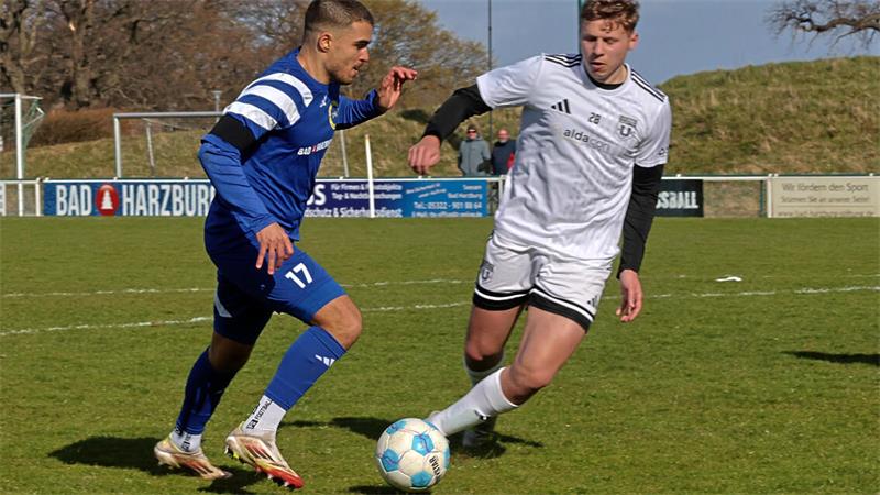 Zwei Fußballspieler in Aktion auf dem Spielfeld, einer in blauem Trikot mit der Nummer 17, der andere in weißem Trikot, beide schauen konzentriert auf den Ball am Boden