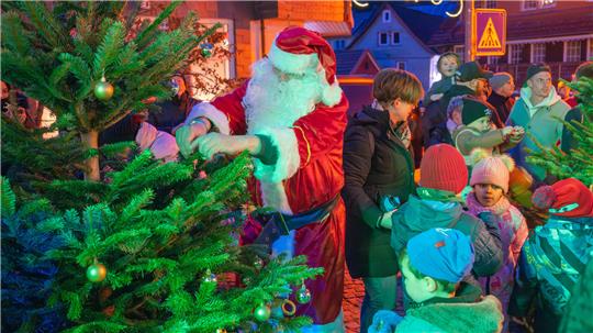 Ein Mann im Weihnachtskostüm schmückt einen Tanne, während hinter ihm viele Kinder stehen.