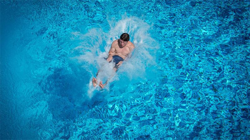 Das Bild zeigt einen jungen Mann, der rückwärts im Wasser eines Swimmingpools einschlägt. 