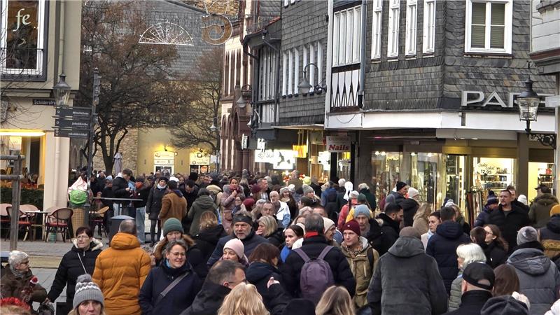 Menschenmenge auf einer belebten Einkaufsstraße mit Fachwerkhäusern und Geschäften, einige tragen Winterkleidung und Rucksäcke.