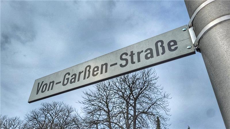 Straßenschild mit der Aufschrift 'Von-Garßen-Straße' an einem Metallpfosten vor bewölktem Himmel und kahlen Bäumen