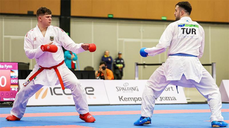 Zwei Karateka im weißen Anzug stehen sich kampfbereit auf einer Matte gegenüber, beide tragen Handschuhe und Gürtel.