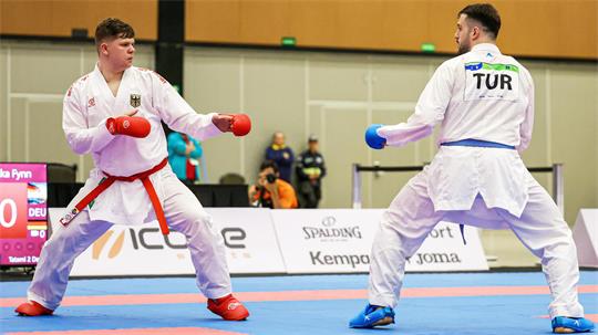 Zwei Karateka im weißen Anzug stehen sich kampfbereit auf einer Matte gegenüber, beide tragen Handschuhe und Gürtel.