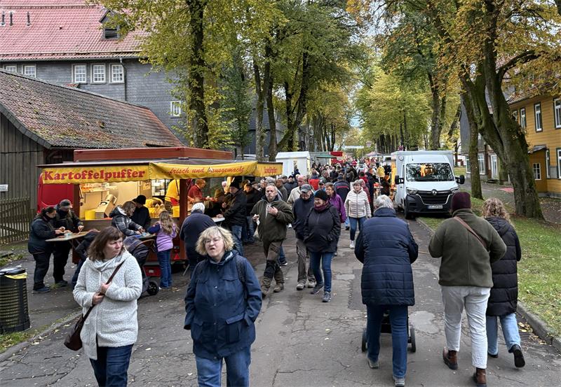 Menschenmenge auf einem von Bäumen gesäumten Weg mit einem gelben Verkaufsstand für Kartoffelpuffer und mehreren Ständen im Hintergrund