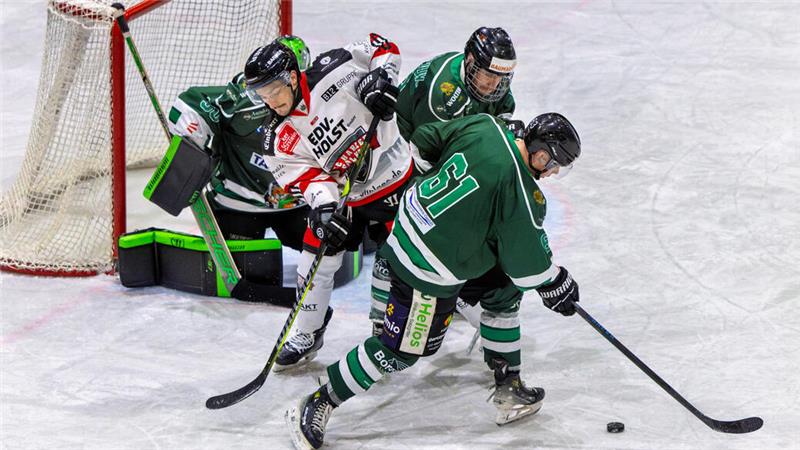 Drei Eishockeyspieler kämpfen vor dem Tor um den Puck, zwei in grüner und einer in weißer Spielkleidung.