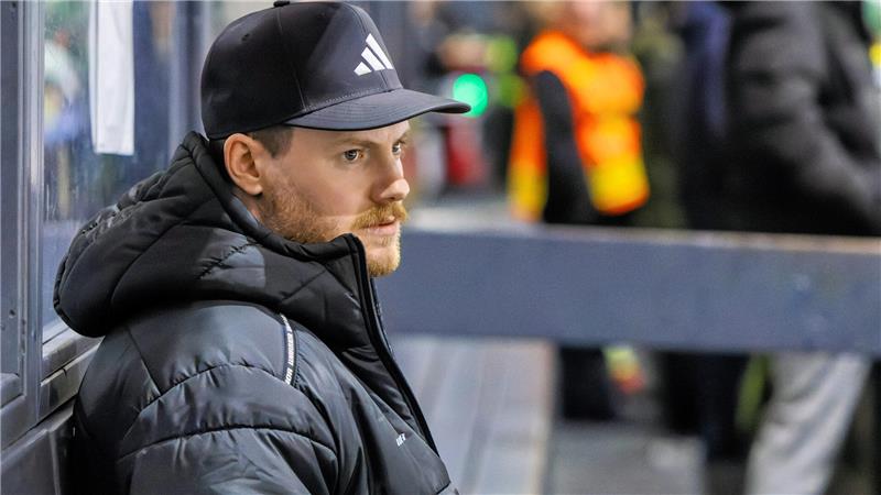 Mann mit schwarzer Adidas-Kappe und schwarzer Steppjacke lehnt an einer Glaswand, im Hintergrund unscharfe Personen in orangefarbenen Warnwesten.