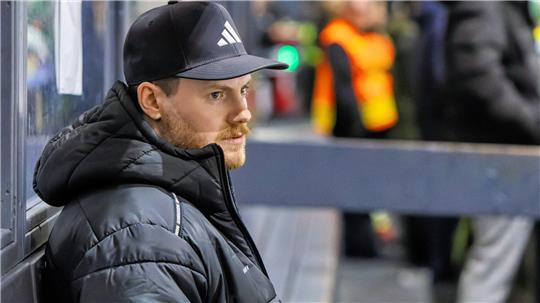 Mann mit schwarzer Adidas-Kappe und schwarzer Steppjacke lehnt an einer Glaswand, im Hintergrund unscharfe Personen in orangefarbenen Warnwesten.