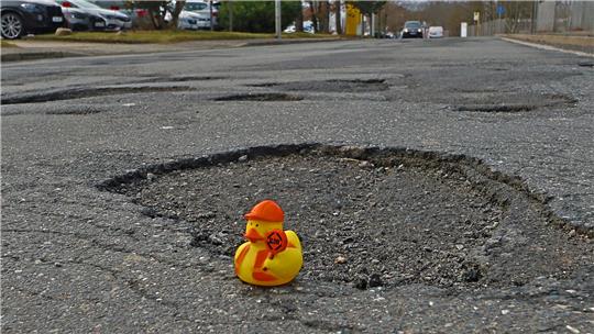 Gummiente mit orangefarbenem Helm und rotem Aufdruck sitzt in einer großen Schlaglochvertiefung auf einer Asphaltstraße.