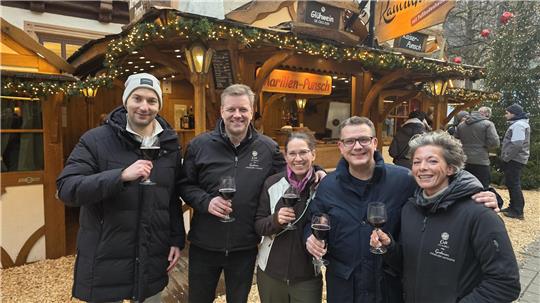Fünf Personen stehen vor einem weihnachtlich geschmückten Holzstand, halten Weingläser und tragen Winterkleidung.
