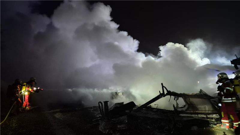 Rauch steigt über Trümmern einer Campingplatzparzelle auf. Feuerwehrleute mit Atemschutzgeräten löschen Glutnester auf dem Gelände.