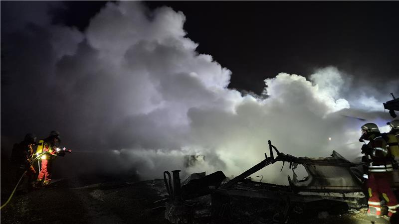 Nach Brand auf Campingplatz: Luftaufnahme zeigt Ausmaß der Zerstörung Rauch steigt über Trümmern einer Campingplatzparzelle auf. Feuerwehrleute mit Atemschutzgeräten löschen Glutnester auf dem Gelände.