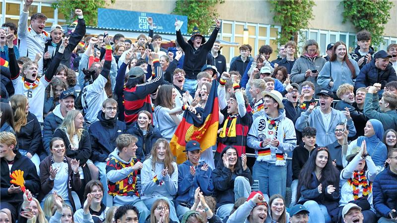 Große jubelnde Menschenmenge mit einigen Personen, die Deutschlandfahnen und -schals tragen, vor einem Gebäude mit gelben Wänden und blauen Schildern