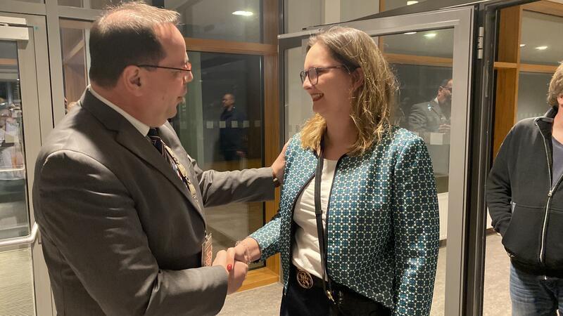 Ein Mann im Anzug und eine Frau mit gemustertem Blazer schütteln sich die Hand in einem Raum mit großen Fenstern.