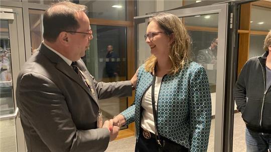 Ein Mann im Anzug und eine Frau mit gemustertem Blazer schütteln sich die Hand in einem Raum mit großen Fenstern.