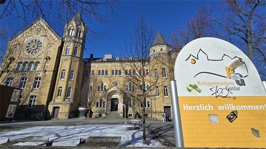 Historisches Schulgebäude mit Türmen und Rosettenfenster im Hintergrund und einem großen Willkommensschild des Ratsgymnasiums Goslar im Vordergrund