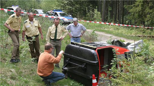 Fünf Männer, darunter Polizisten in Uniform, stehen an einem umgekippten roten Fahrzeug neben einem Waldweg, der mit Absperrband gesichert ist.