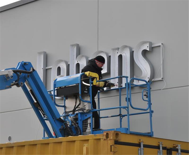 Ein Arbeiter auf einer Hebebühne montiert silberne Buchstaben an grauer Fassade eines Gebäudes.