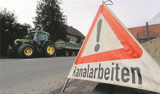 Ein Aufsteller mit der Aufschrift Kanalarbeiten steht am Straßenrand, im Hintergrund fährt ein Trecker mit Anhänger.