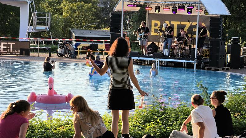 Menschen sitzen und stehen am Ufer eines Schwimmbeckens, im Wasser schwimmt ein aufblasbarer Flamingo, im Hintergrund spielt eine Band auf einer Bühne mit Überdachung