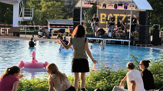 Menschen sitzen und stehen am Ufer eines Schwimmbeckens, im Wasser schwimmt ein aufblasbarer Flamingo, im Hintergrund spielt eine Band auf einer Bühne mit Überdachung