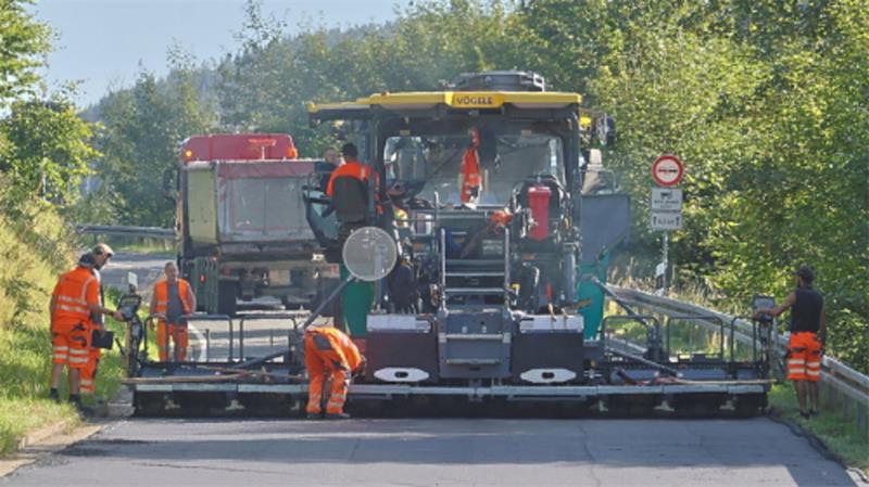 Straßenbaumaschine mit mehreren Arbeitern in orangefarbener Schutzkleidung bei der Asphaltierung einer Straße in ländlicher Umgebung