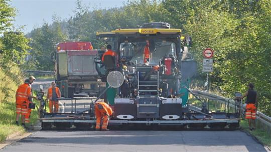 Straßenbaumaschine mit mehreren Arbeitern in orangefarbener Schutzkleidung bei der Asphaltierung einer Straße in ländlicher Umgebung
