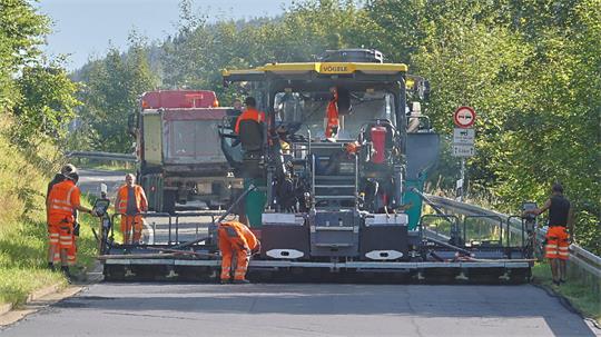 Straßenbaumaschine mit mehreren Arbeitern in orangefarbener Schutzkleidung bei der Asphaltierung einer Straße in ländlicher Umgebung