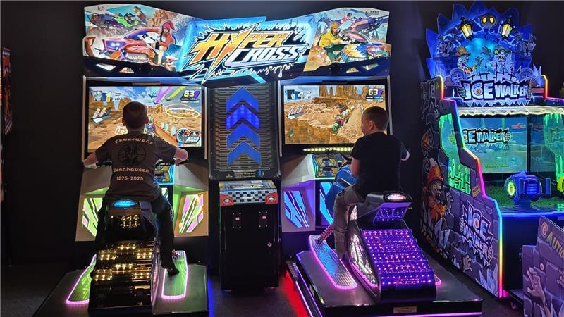 Zwei Personen sitzen auf beleuchteten Motorradsimulatoren vor Bildschirmen mit Rennspielgrafiken in einer Spielhalle.