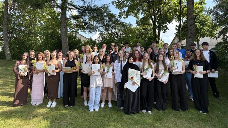 Gruppe von jungen Menschen steht im Freien auf Grasfläche vor Bäumen, viele halten Urkunden und Blumen.