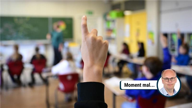 Nahaufnahme einer Hand, die im Klassenzimmer zum Melden erhoben ist, im Hintergrund unscharfe Schüler und Lehrkraft