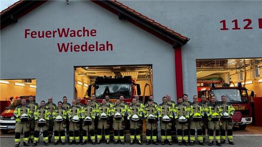 Feuerwehrleute in Schutzkleidung stehen in einer Reihe vor der Feuerwache Wiedelah mit geöffneten Toren und sichtbaren Einsatzfahrzeugen