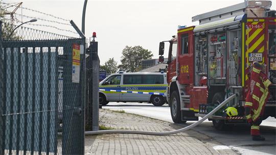 Ein Polizeifahrzeug, sowie ein Feuerwehrlöschfahrzeug stehen vor einem Zaun eines Firmengeländes.