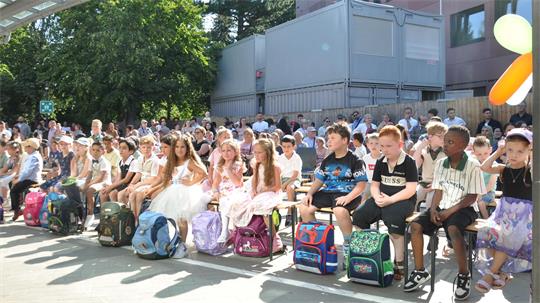 Gruppe von Kindern sitzt in einer Reihe auf Stühlen im Freien mit Schulranzen vor sich und Zuschauern im Hintergrund