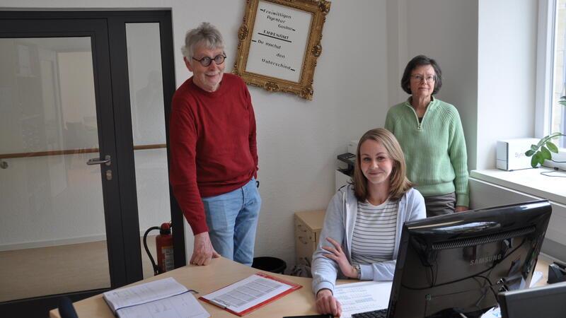 Drei Personen in einem Büro, eine sitzt am Schreibtisch vor einem Computer, zwei stehen daneben, im Hintergrund ein Fenster und eine Tür