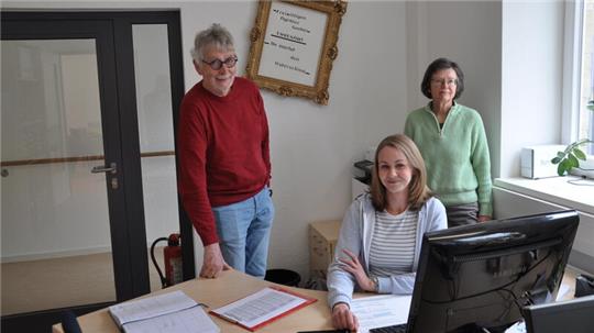 Drei Personen in einem Büro, eine sitzt am Schreibtisch vor einem Computer, zwei stehen daneben, im Hintergrund ein Fenster und eine Tür