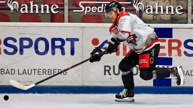 Eishockeyspieler in weißem Trikot und schwarzer Hose schießt Puck auf Eisfläche vor Werbebanden mit roter Zuschauertribüne im Hintergrund