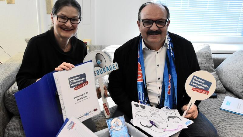 Zwei Personen sitzen auf einem Sofa, eine hält einen blauen Ordner mit der Aufschrift 'Zukunft jetzt gestalten', die andere trägt einen Schal und hält ein rundes Schild mit der Aufschrift 'Deutscher Präventionstag 2023'.