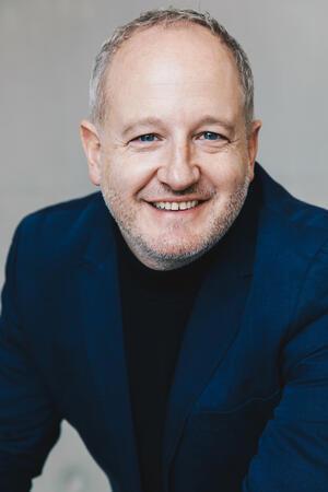 Gandersheimer Domfestspiele räumen bei Musical-Award ab Mann mit grauem Bart trägt schwarzen Rollkragenpullover und dunkelblauen Blazer vor grauem Hintergrund.