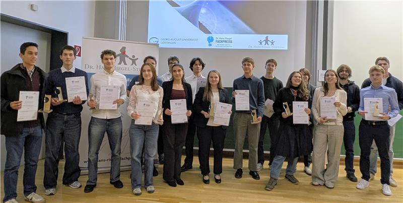 Gruppe von Menschen steht in einem Saal vor einer Leinwand, die Text zur Verleihung des Dr.-Hans-Riegel-Fachpreises zeigt, viele halten Urkunden in der Hand.