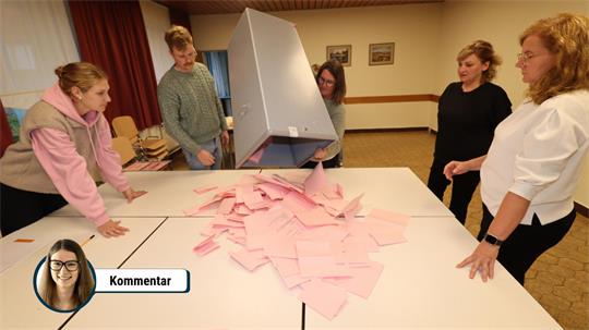 Fünf Frauen kippen einen großen Behälter mit vielen rosa Zetteln auf einen Tisch in einem Raum mit Teppichboden und Bildern an der Wand.